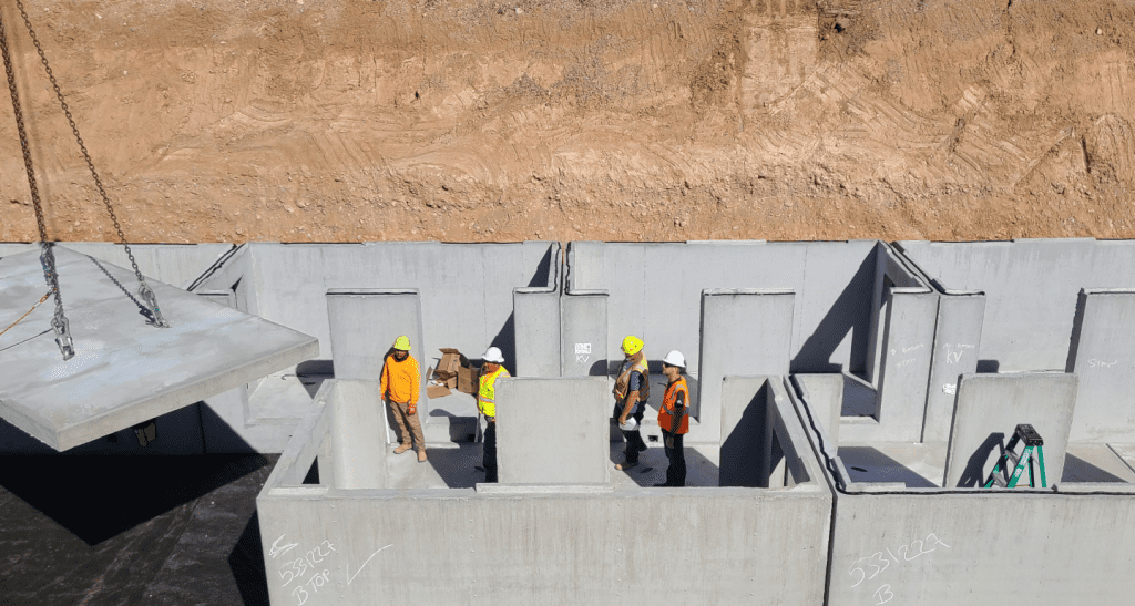 Construction workers in safety vests and helmets stand inside a partially built concrete structure at a worksite, while a large concrete slab is being lowered by a crane.